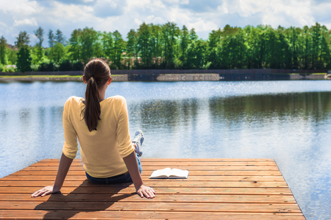 Woman sitting on a wooden dock looking at a lake with an open book beside her